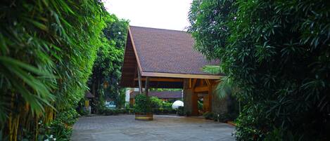 A building with a sloped roof and a covered entrance area, surrounded by lush greenery.