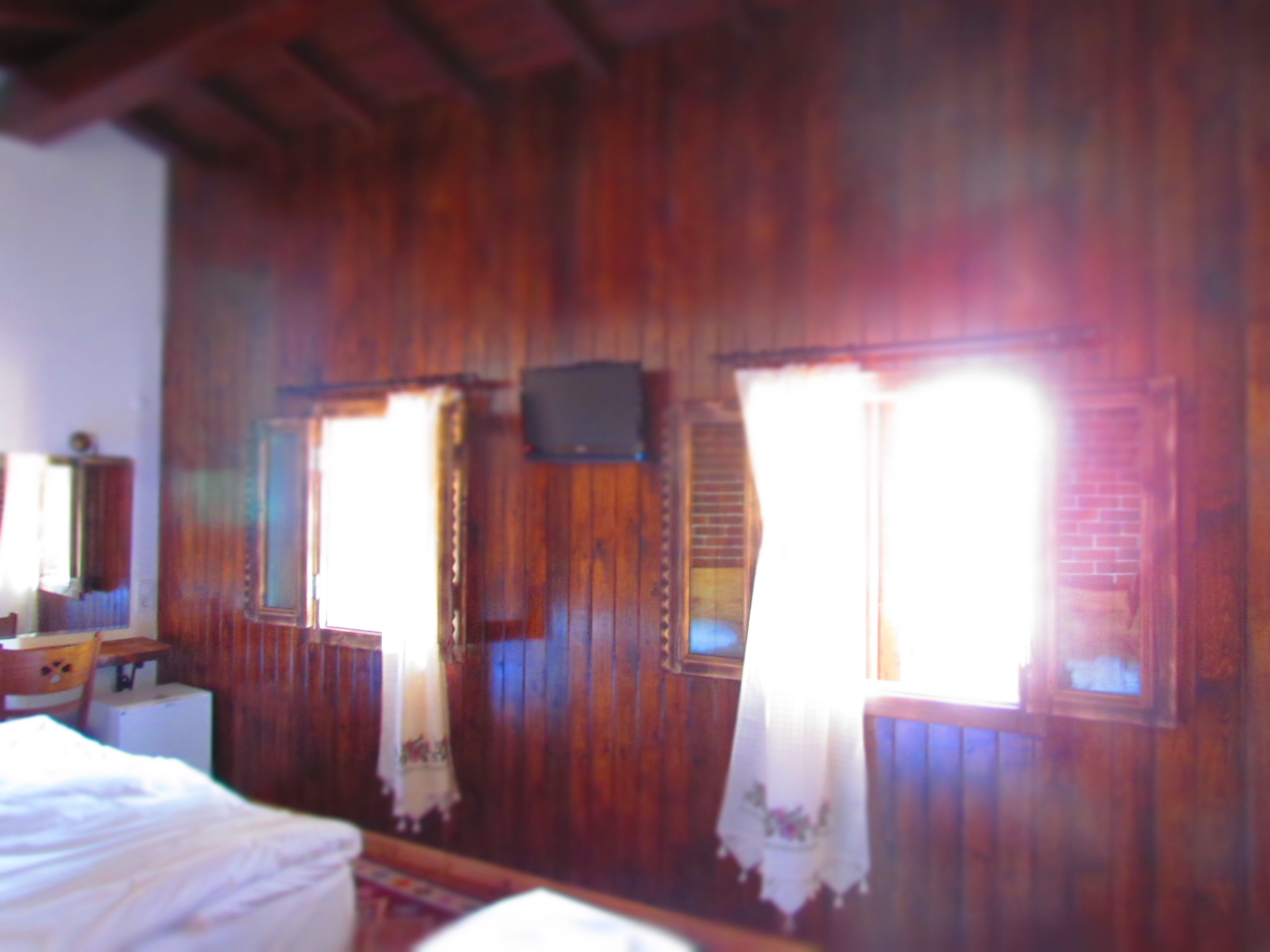 Ein Zimmer mit Holzwänden, einem Bett, einem kleinen Tisch und zwei mit Vorhängen versehenen Fenstern.
