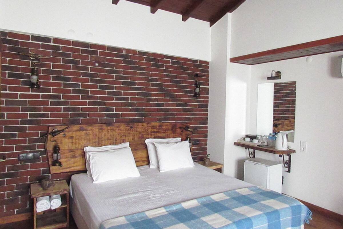 Un dormitorio con pared de ladrillo visto, cabecero de madera, cama con ropa de cama blanca, manta azul y blanca a cuadros, mesita de noche con lámpara y un estante pequeño con artículos de aseo.