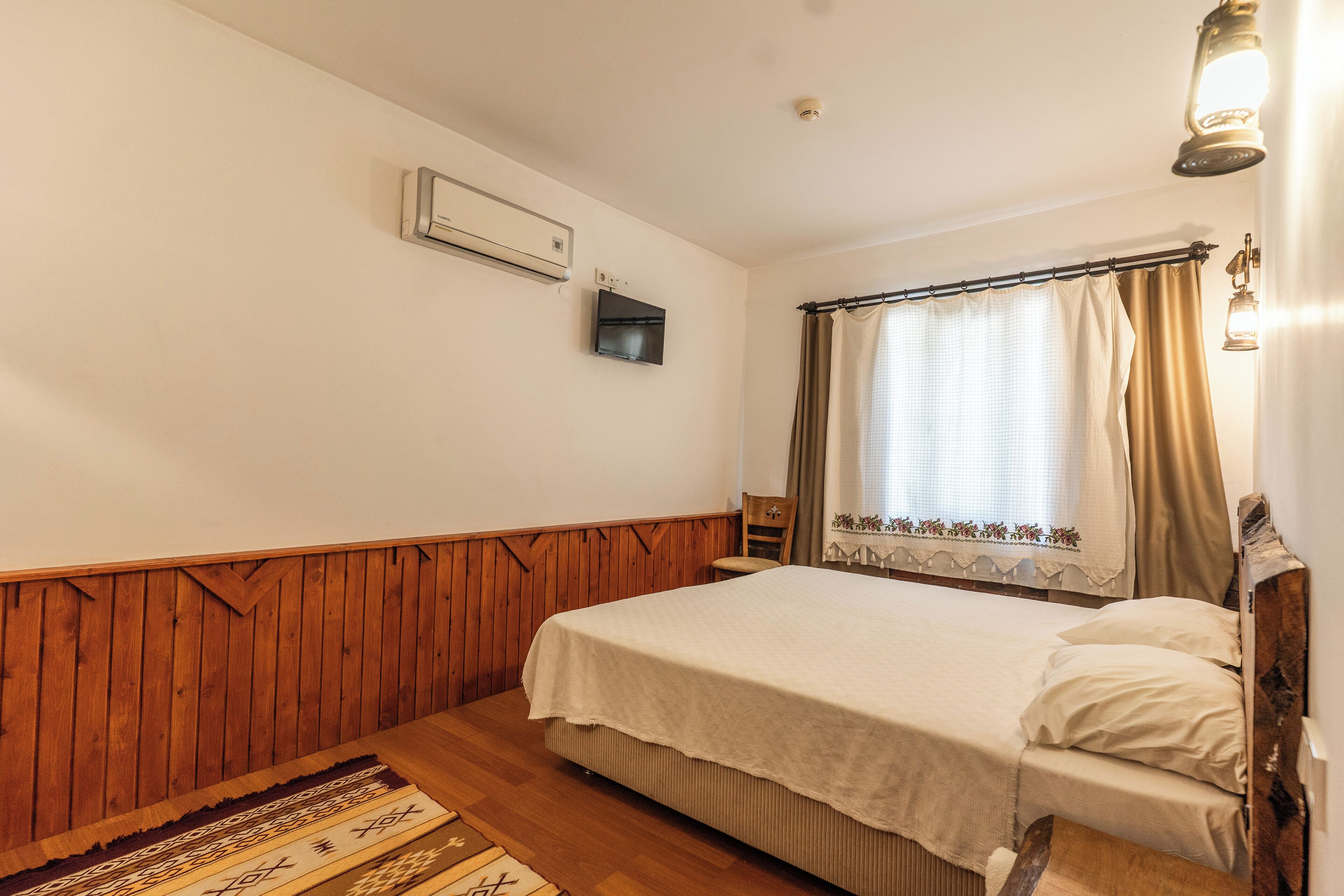 A bedroom with a wooden headboard, a bed with a white bedspread, a wooden floor, a wall-mounted air conditioner, a television, and a window with curtains.