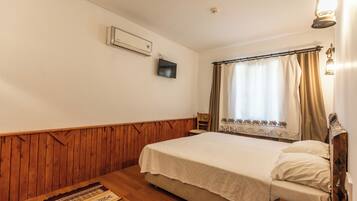 A bedroom with a wooden headboard, a bed with a white bedspread, a wooden floor, a wall-mounted air conditioner, a television, and a window with curtains.