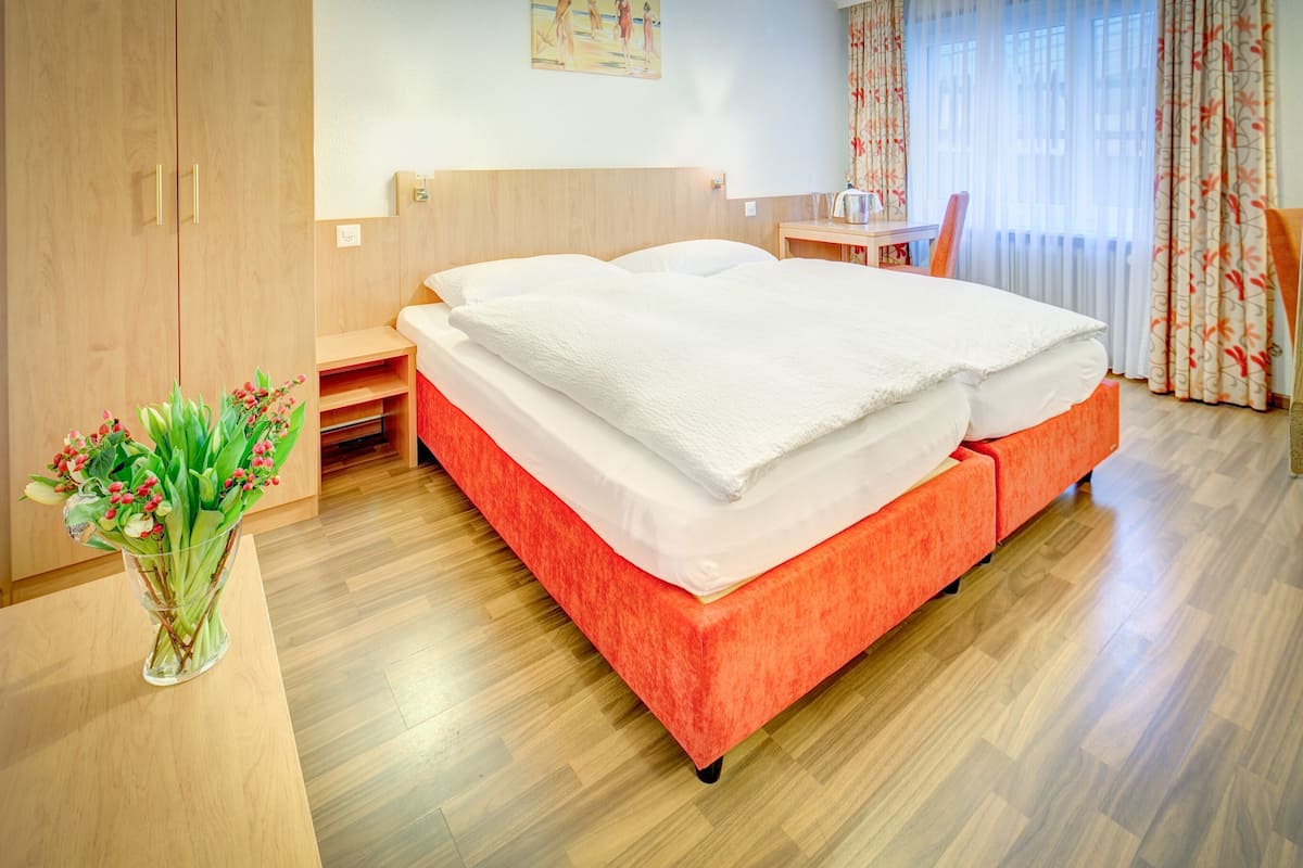 A hotel room with a bed, a wooden wardrobe, a small table with a chair, and a vase of flowers.