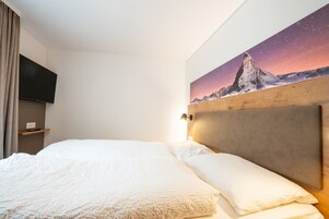 Una habitación de hotel con cama, televisor montado, un cuadro de una montaña y cabecero de madera.