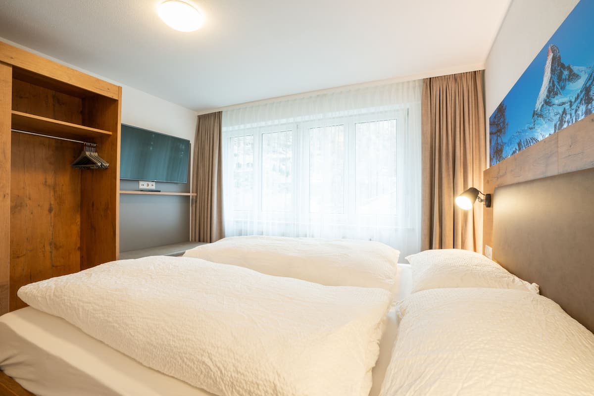 A hotel room with two beds, a wooden wardrobe, a shelf with hangers, a window with curtains, and a painting on the wall.