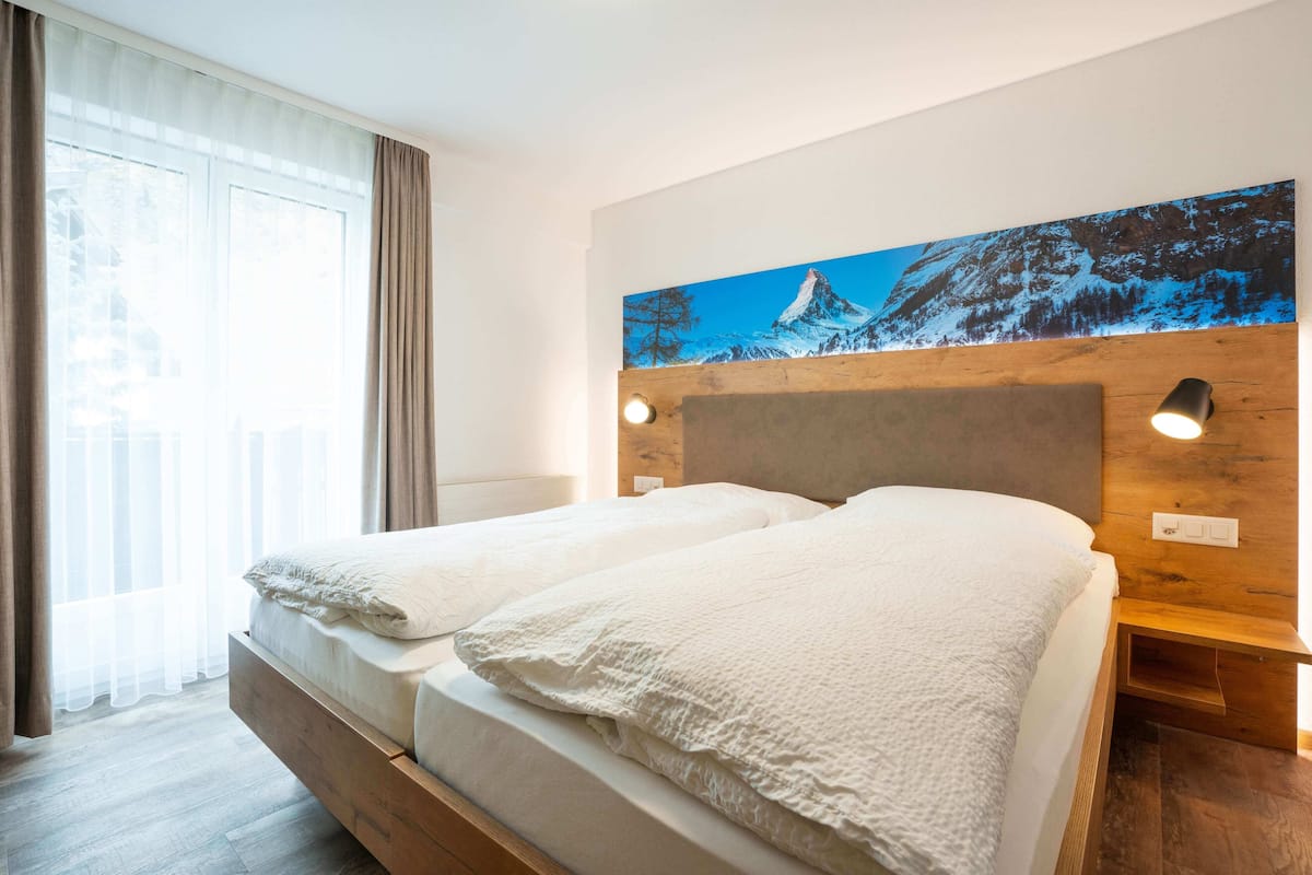 A double bed with a wooden headboard, two bedside tables with lamps, a large window with sheer curtains, and a painting of a mountain landscape on the wall.