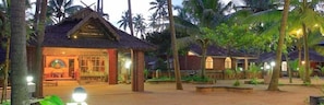 A resort with thatched-roof buildings, palm trees, and a well-lit entrance. - Cherai Beach Resort (Kochi)