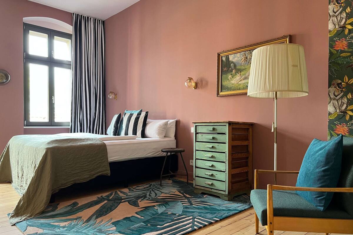 A bedroom with a bed, a dresser, a chair, a lamp, a window with curtains, and a painting on the wall.