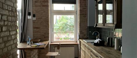 A kitchen with a large window, wooden cabinets, a radiator, and a small table with a chair.