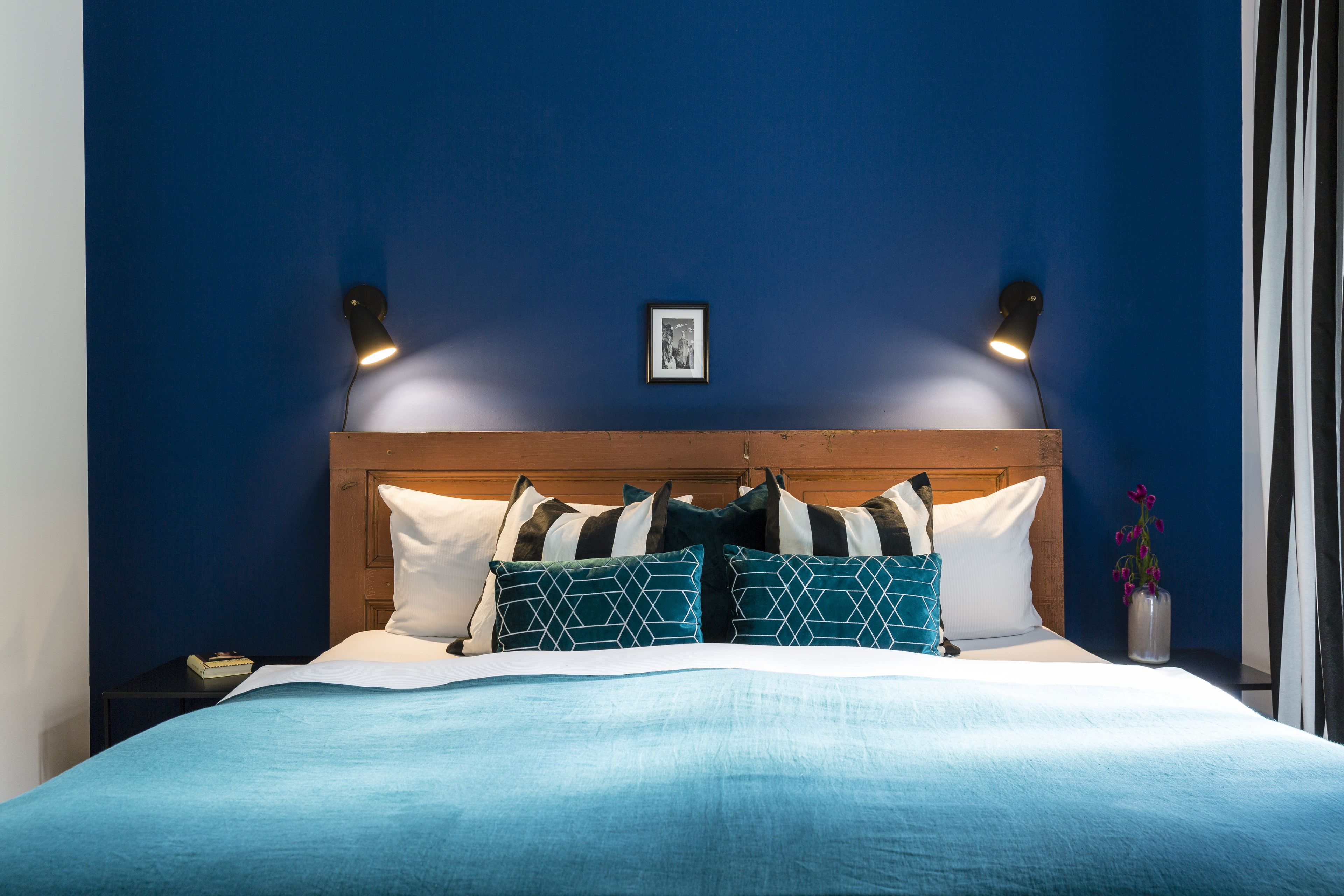 A neatly made bed with a blue bedspread and multiple pillows, two wall-mounted reading lamps, and a small framed picture on the wall.