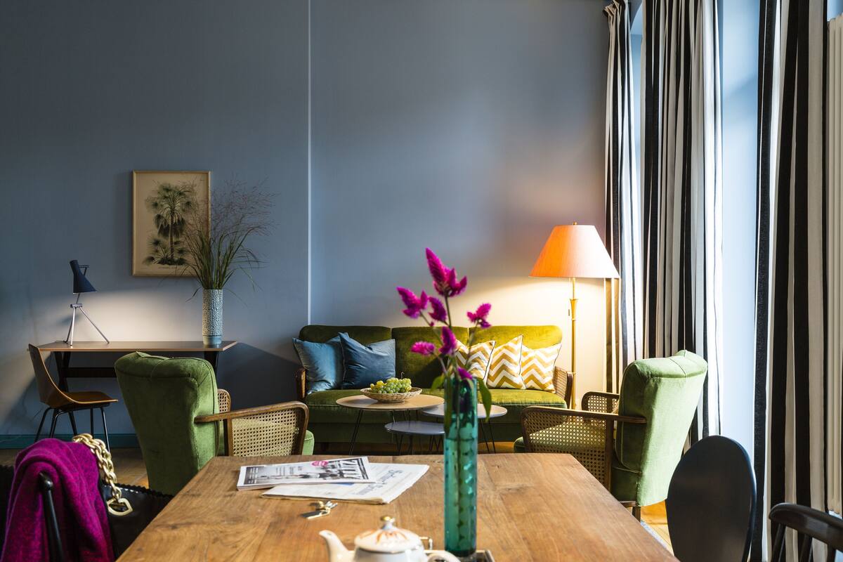A dining area with a wooden table, chairs, a vase with flowers, a lamp, and a sofa with cushions.