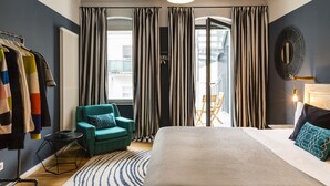 Una habitación de hotel moderna con una cama grande, un sillón verde azulado, una mesa redonda y vistas de la ciudad a través del ventanal.