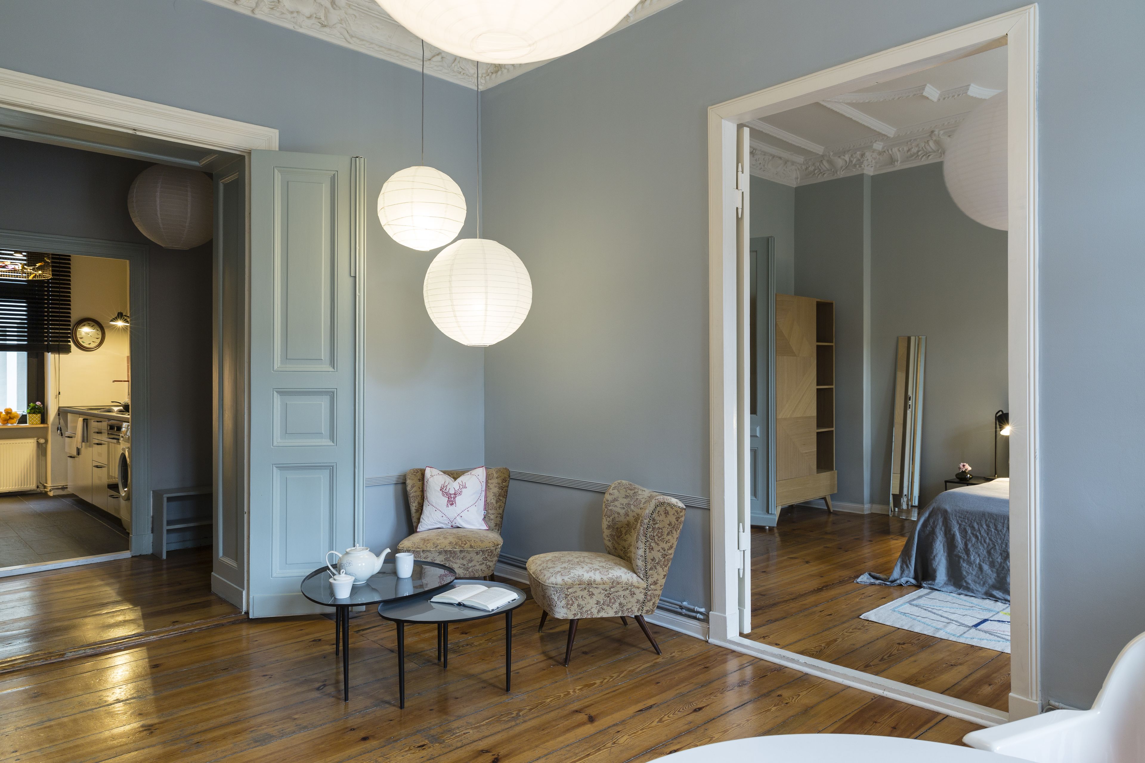 A modern living room with a wooden floor, a round coffee table, two armchairs, and a large mirror reflecting a bedroom.
