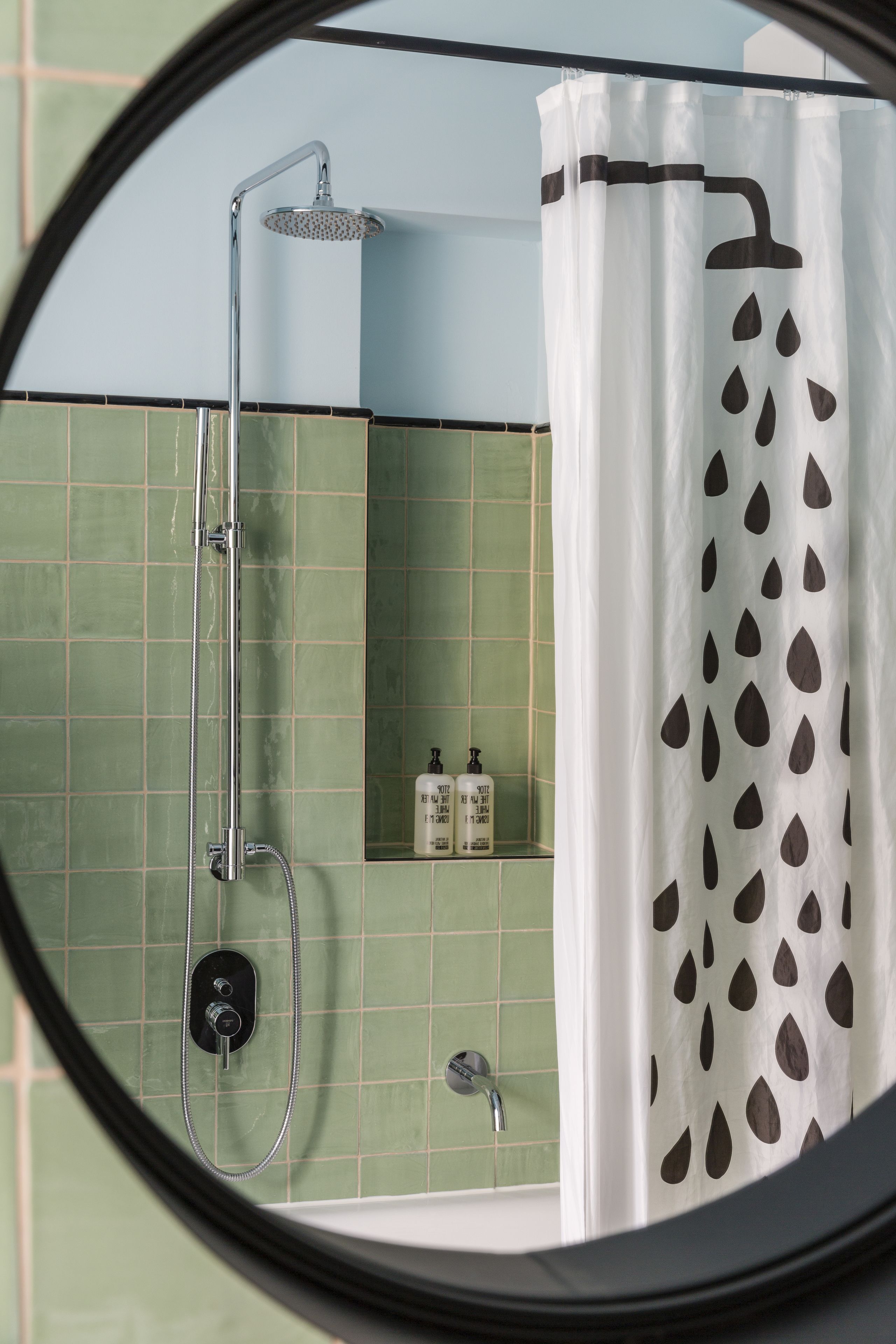 A bathroom with a shower, green tiled walls, and a white shower curtain with black leaf patterns.