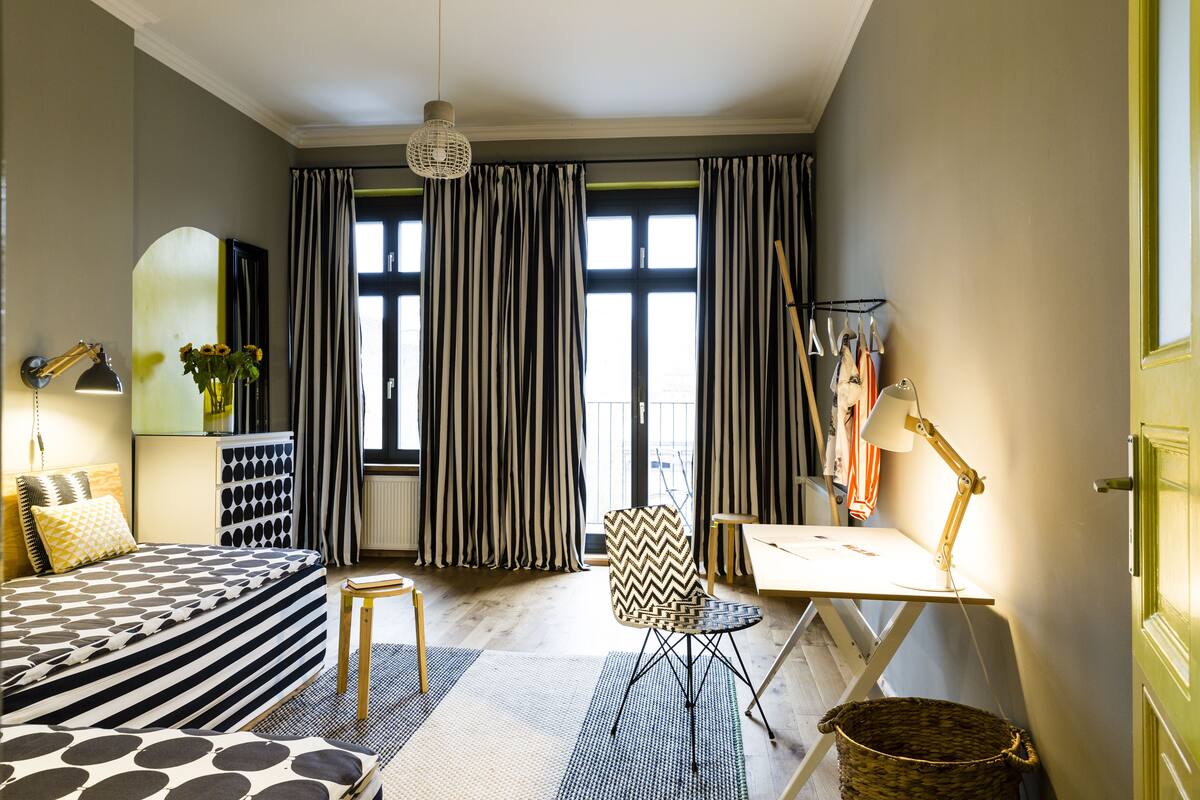 A bedroom with a striped bed, a desk with a lamp, a chair, a hanging lamp, and a large window with curtains.