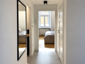 A narrow hallway leading to two bedrooms with a large mirror on the left, a window on the right, and a ceiling light. - Rainbow Apartments 2 (Kraków)
