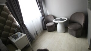 A modern hotel room with a tufted headboard, a small bedside table with a lamp, two armchairs, a round table, and a large window with curtains. - Rainbow Apartments 2 (Kraków)