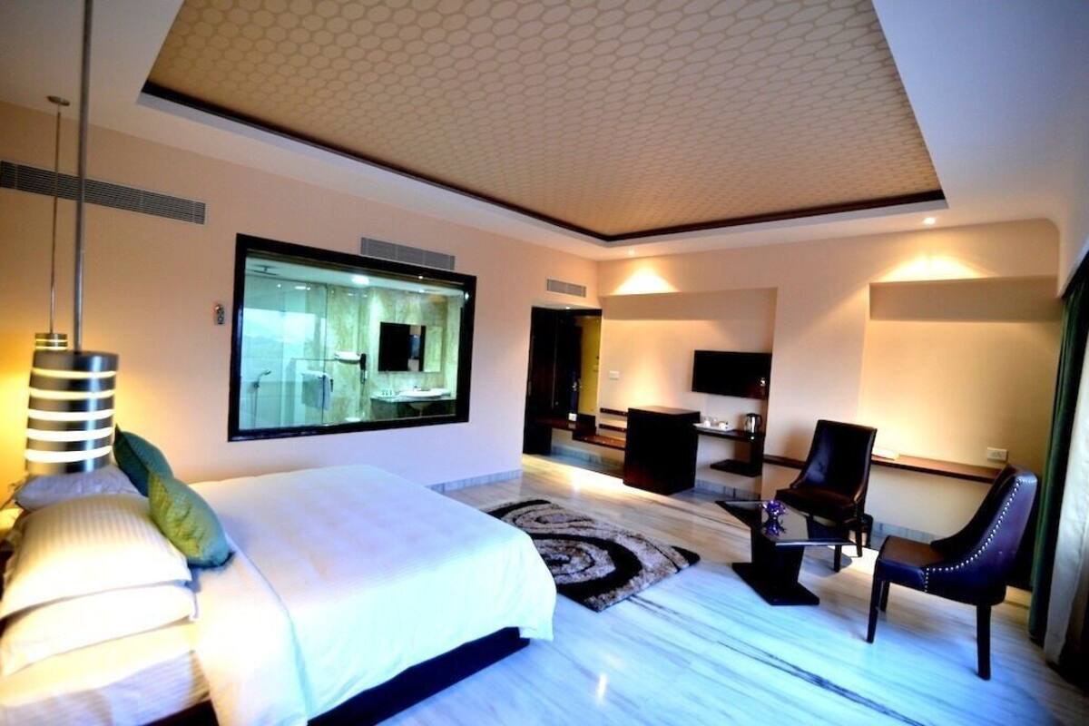 A modern hotel room with a large bed, a sitting area with a chair, a TV, and a bathroom visible through a glass door.