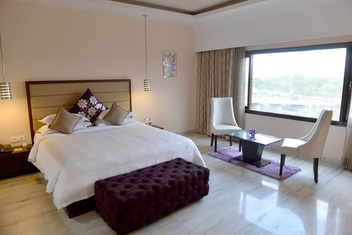 A modern hotel room with a large bed, a sitting area with a table and chairs, and a large window offering a city view.
