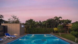 A rectangular swimming pool with a tiled edge, surrounded by a low stone wall and a hedge. There are lounge chairs and a table with a fruit bowl on the left side. - Zone Connect by The Park Udaipur (Udaipur)