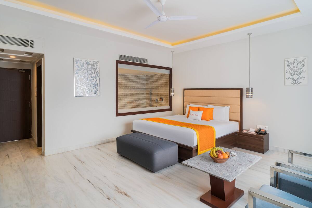 A hotel room with a bed, a desk, a chair, a small table with fruit, a nightstand, and a ceiling fan.
