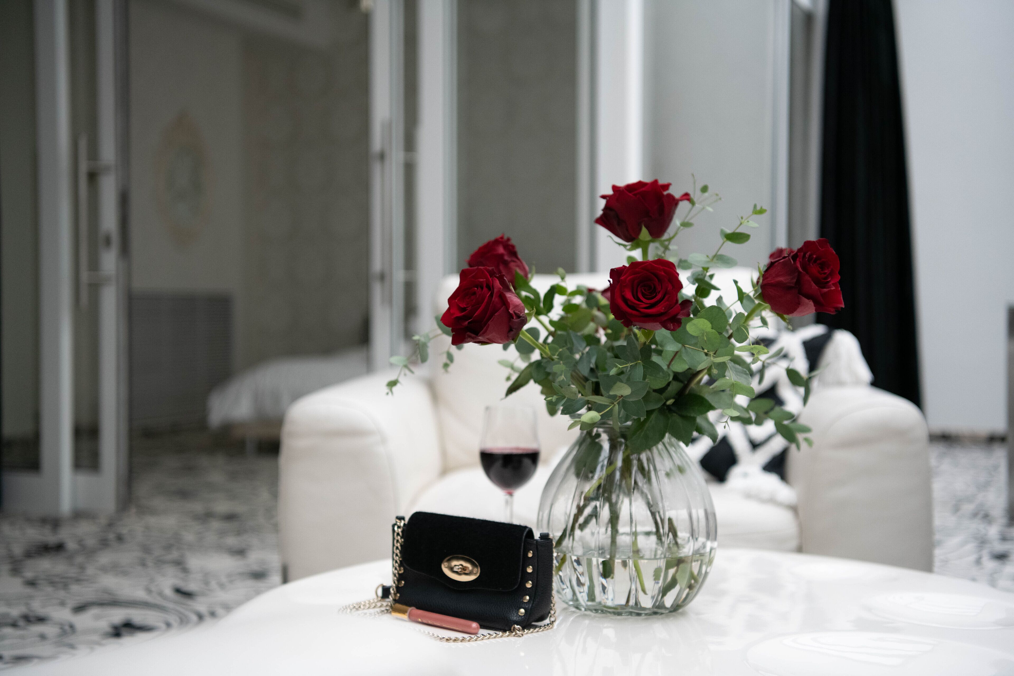 Ein weißes Sofa, eine Glasvase mit roten Rosen, ein Weinglas und eine schwarze Handtasche auf einem weißen Tisch.