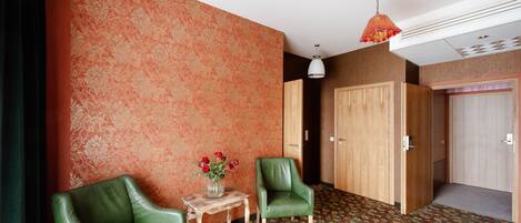 A room with a patterned carpet, two green chairs, a small table with a vase of flowers, and a wooden door.