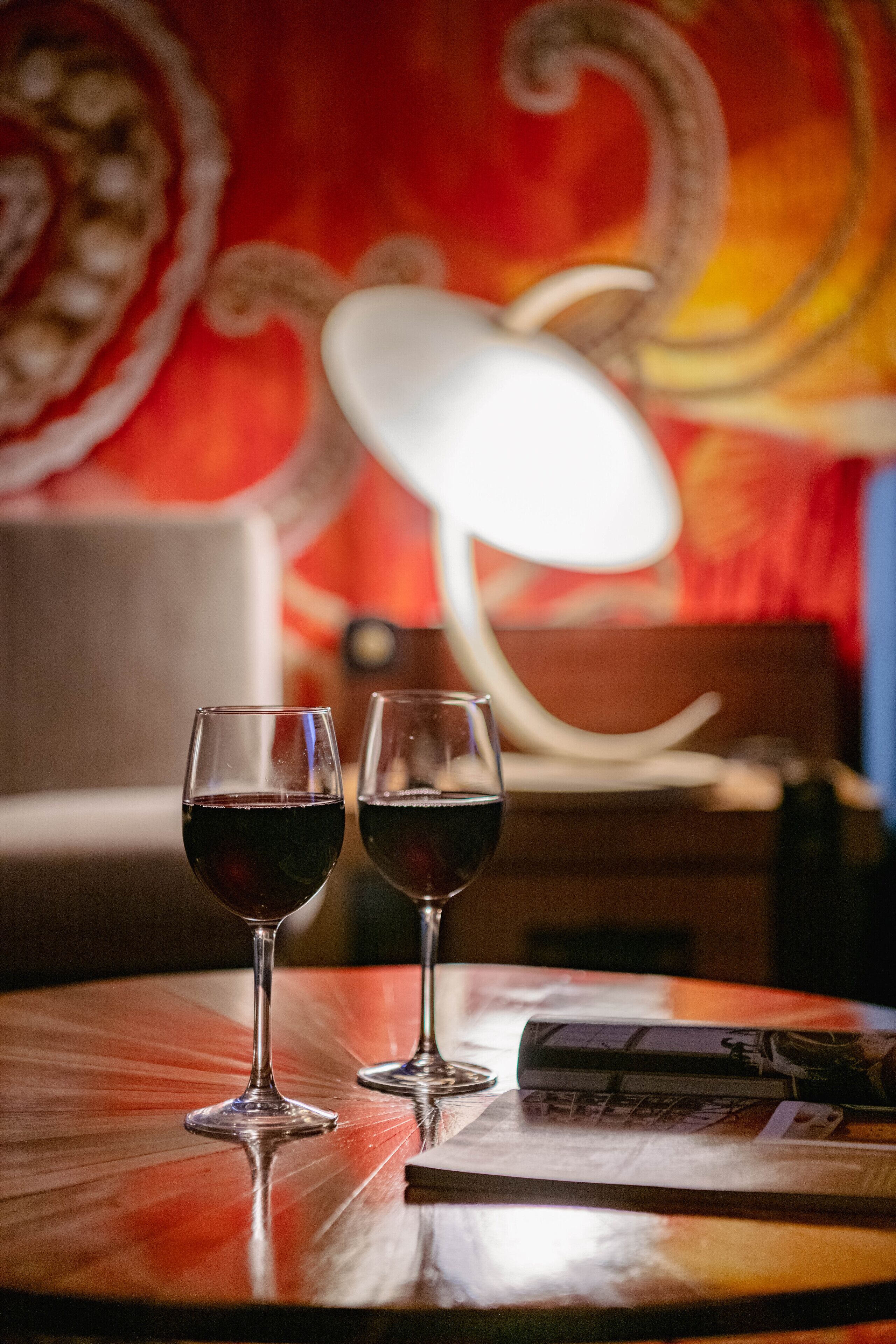 Two wine glasses on a wooden table with a magazine, in a room with a red tapestry and a white lamp in the background.