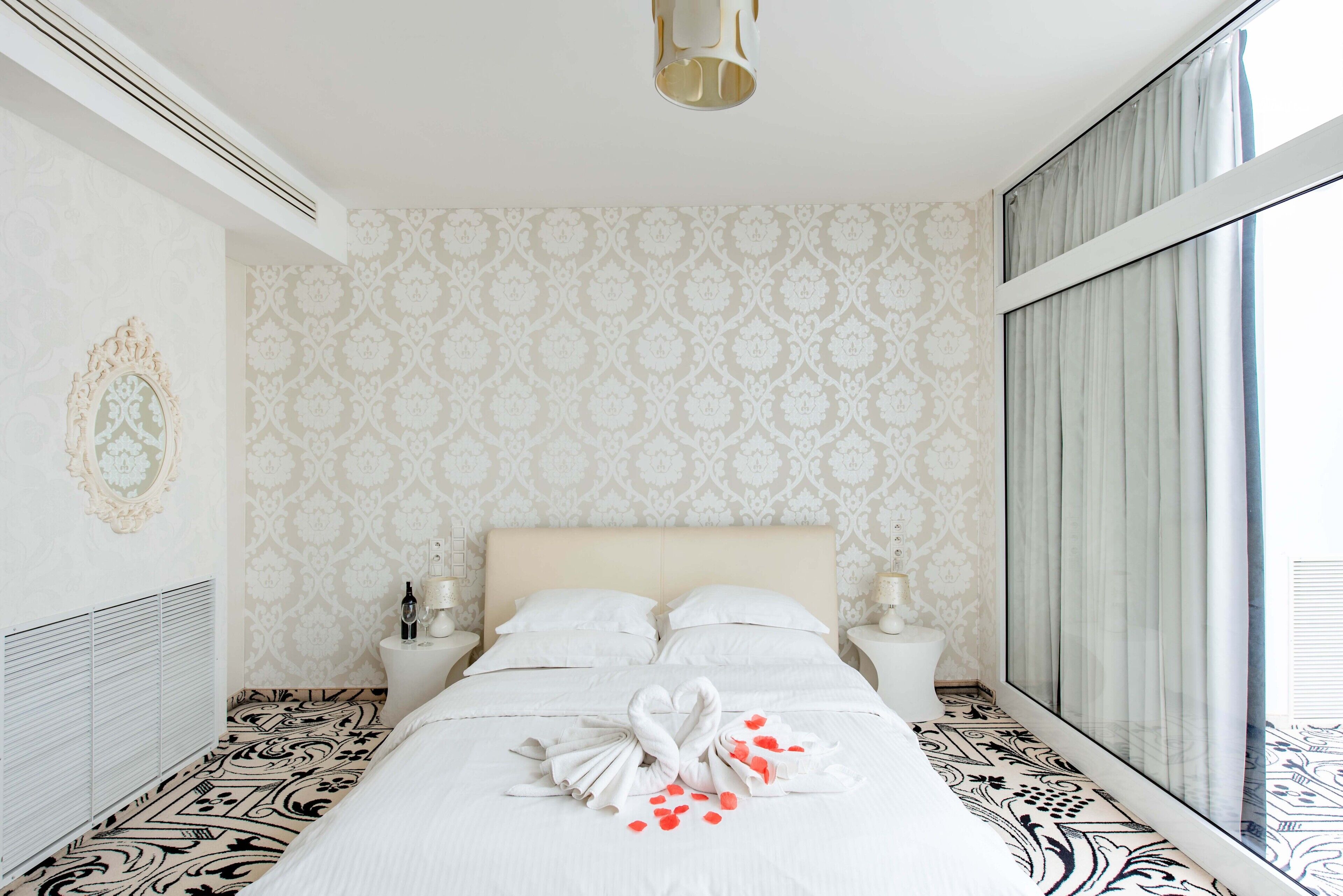 A hotel room with a large bed, two bedside tables, a nightstand, a wall-mounted mirror, and a window with curtains.