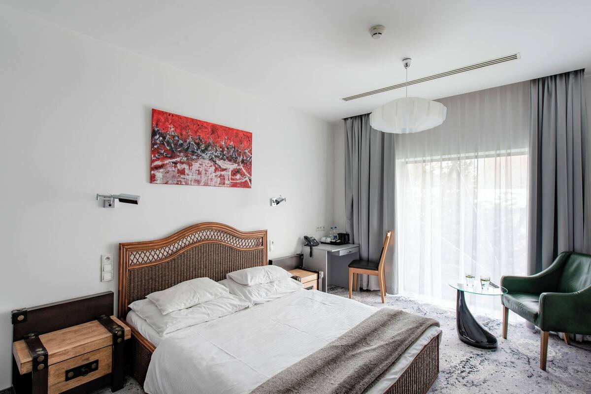 A modern hotel room with a large bed, a desk, a chair, and a view of the outdoors through sheer curtains.