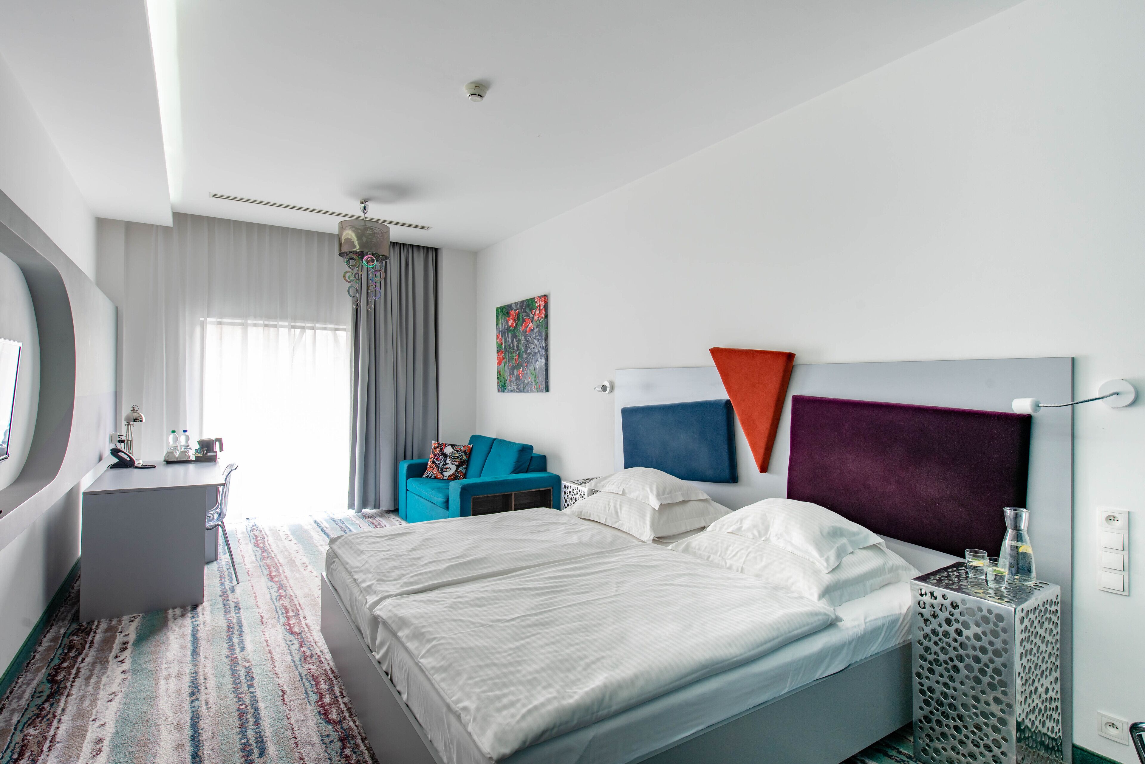 Ein modernes Hotelzimmer mit einem großen Bett, einem Schreibtisch und einem Stuhl. Es gibt ein Fenster mit Vorhängen, ein Bild an der Wand und ein außergewöhnliches Kopfteildesign.