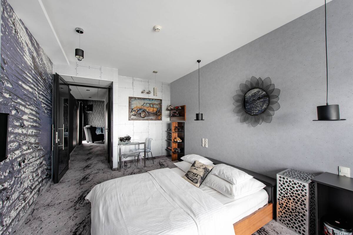 A modern bedroom with a bed, a desk, and a chair. There is a painting on the wall and a decorative item above the bed.