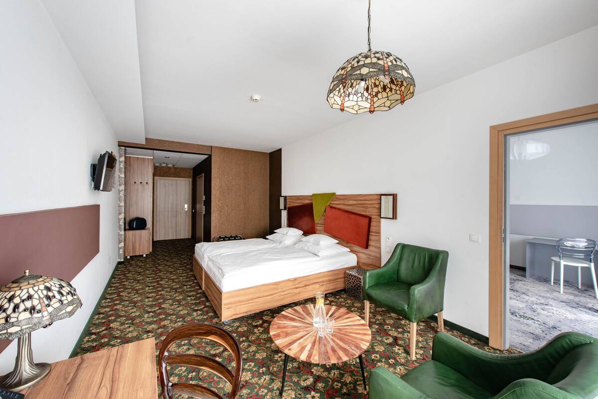 A hotel room with a bed, green chairs, a round wooden table, a lamp, and a hanging light fixture.