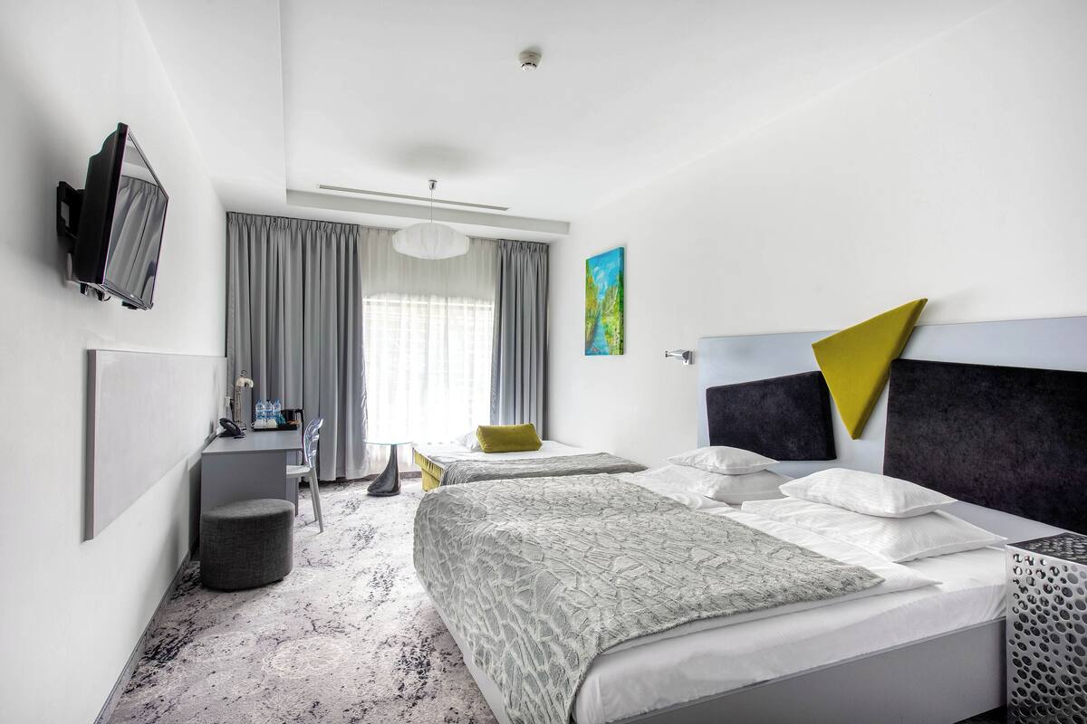 A modern hotel room with two beds, a desk, a chair, a TV, and a large window with curtains.