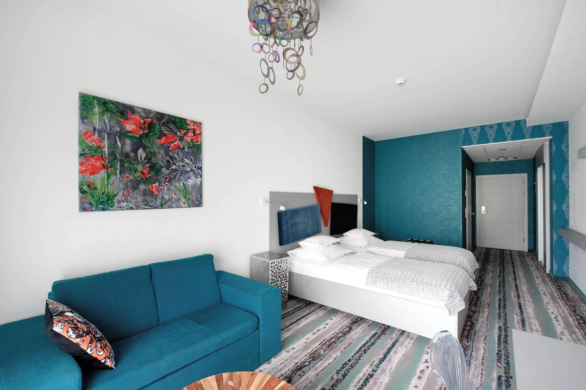 A modern bedroom with a bed, a blue sofa, a colorful abstract painting, and a patterned carpet.