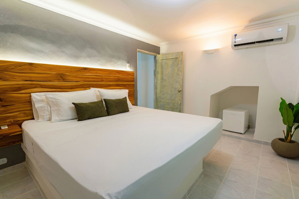 A bedroom with a large bed, a wooden headboard, a small bedside table, a nightstand, a wall-mounted air conditioner, a door, and a potted plant.