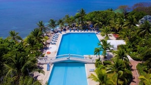Vue aérienne d’un complexe hôtelier doté d’une grande piscine, entourée de palmiers, avec vue sur l’océan.