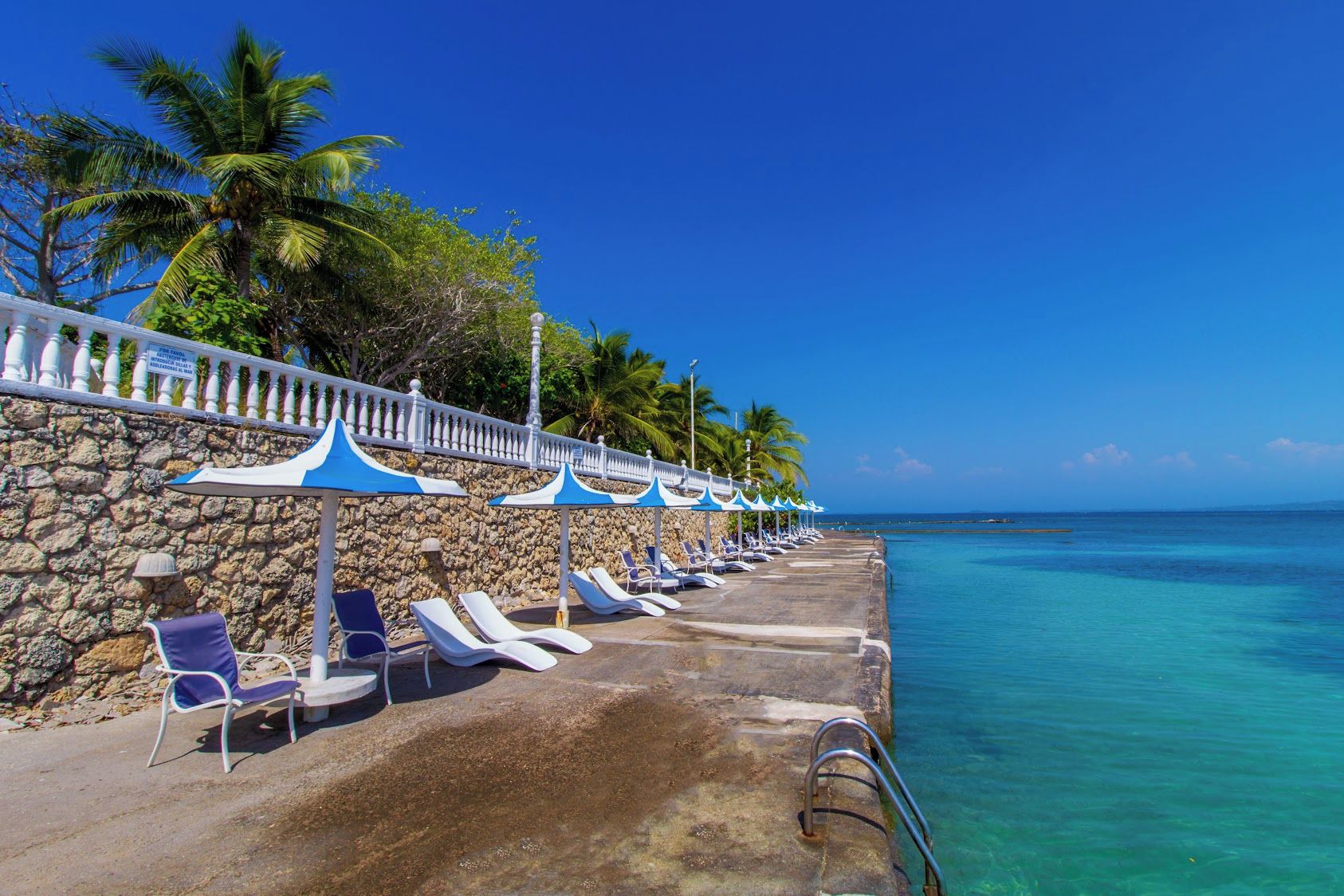 A seaside resort with a row of sun loungers and umbrellas, a stone wall, and a clear view of the ocean.