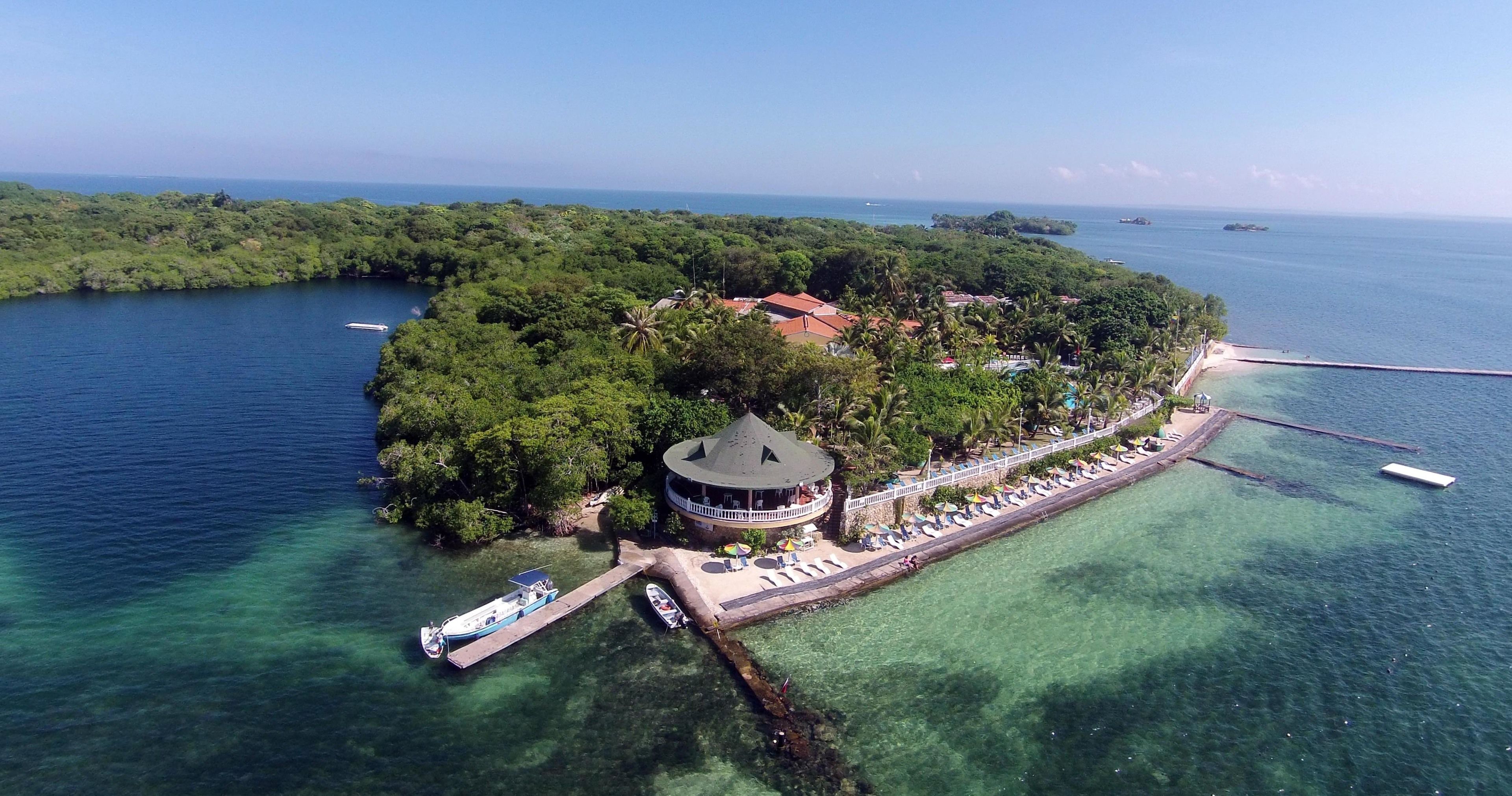 An island with a circular building surrounded by a beach area with sun loungers and a pier.