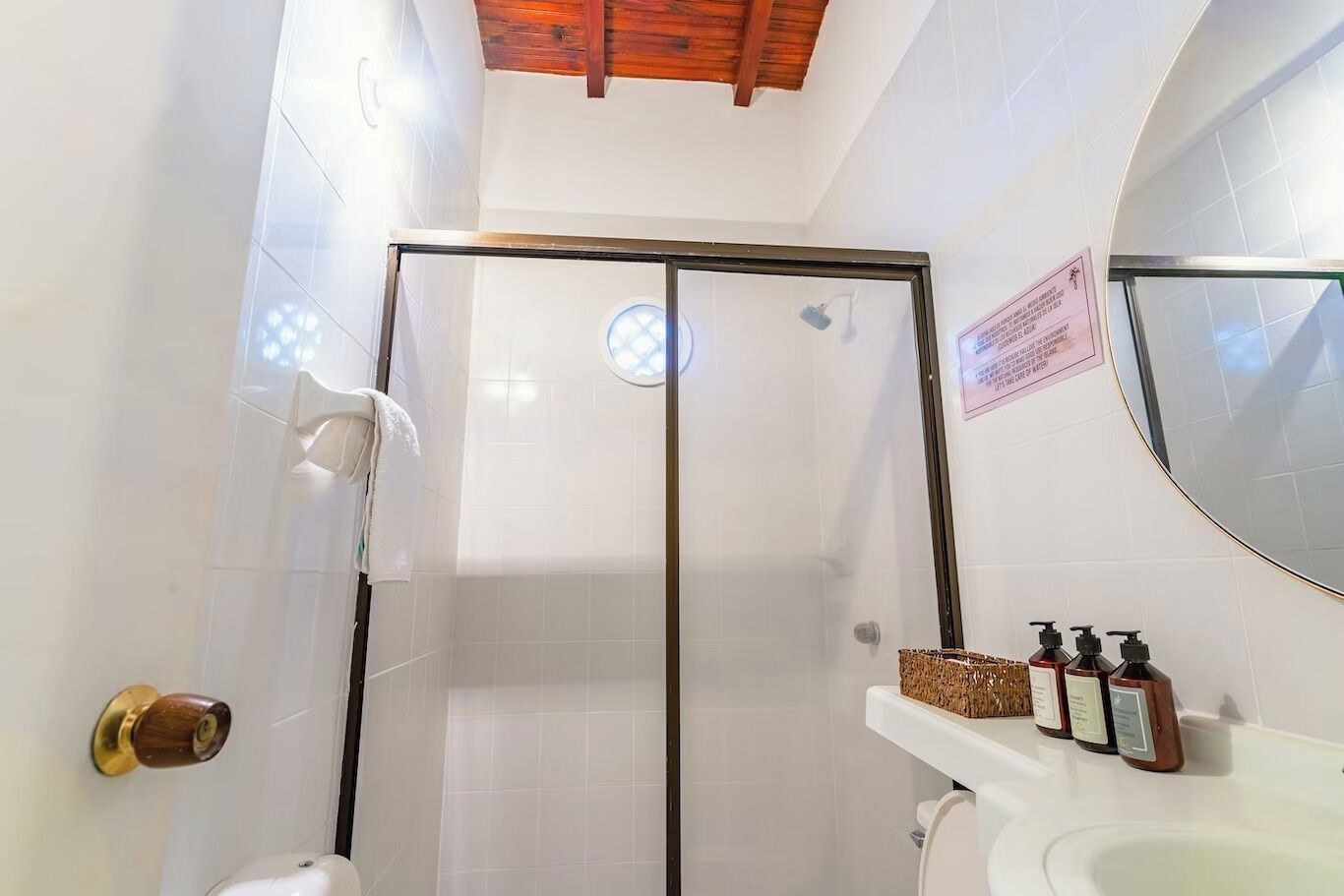 Un baño con mampara de vidrio, un lavamanos blanco con dispensadores de jabón y techo de madera.