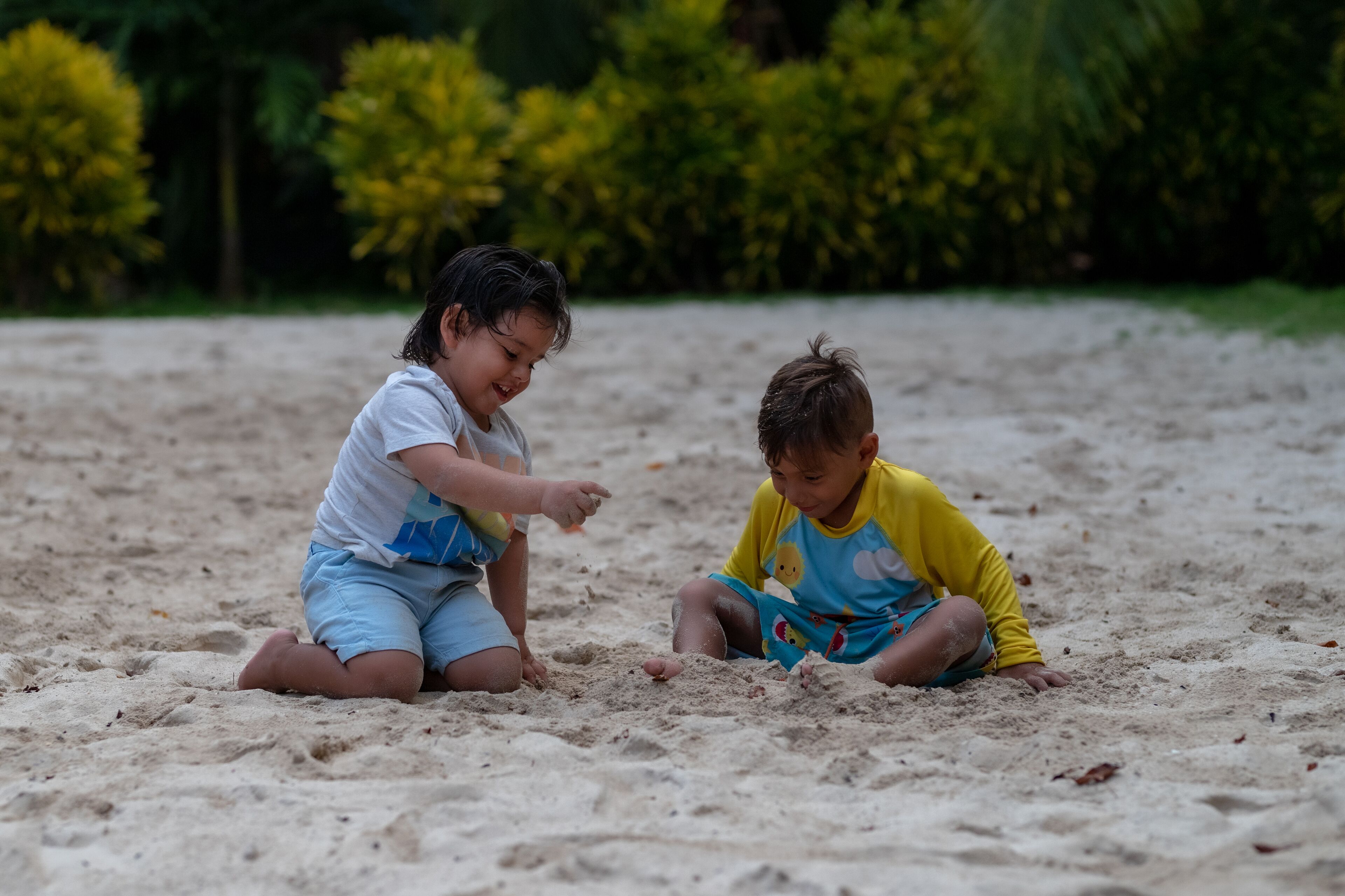 Two children playing on a sandy beach with greenery in the background.
