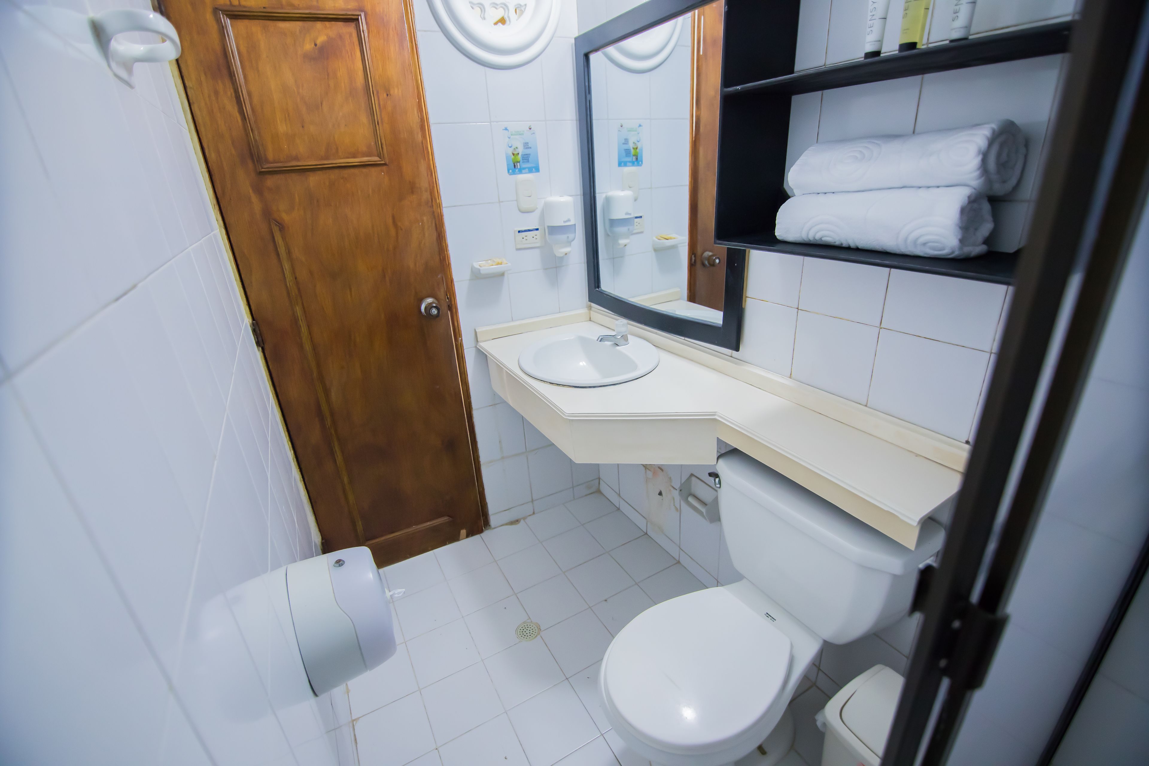 A bathroom with a wooden door, white tiles, a sink, a toilet, a mirror, and shelves with folded towels.