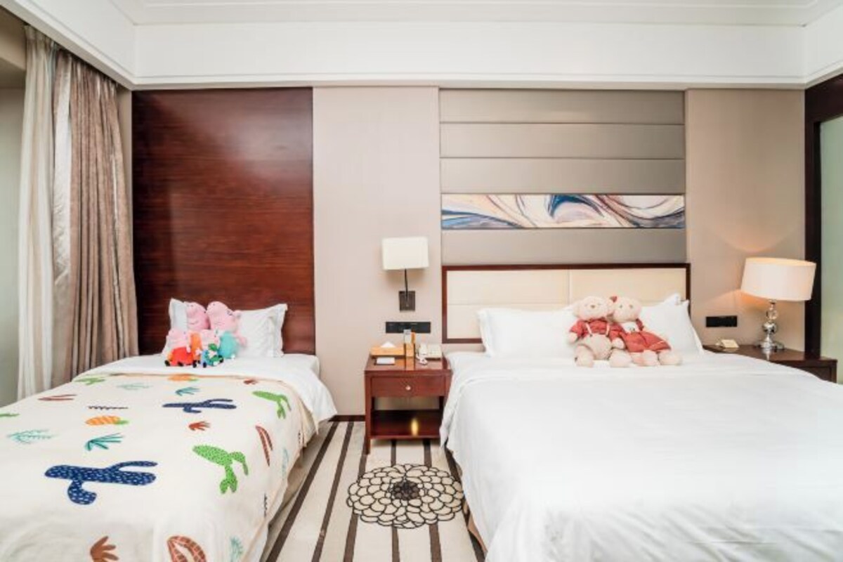 A hotel room with two beds, a wooden headboard, a wall art piece, a nightstand with a lamp, and a decorative rug.