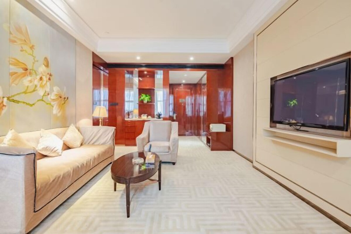 A modern hotel room with a sofa, a coffee table, a TV mounted on the wall, and a large window with a view.