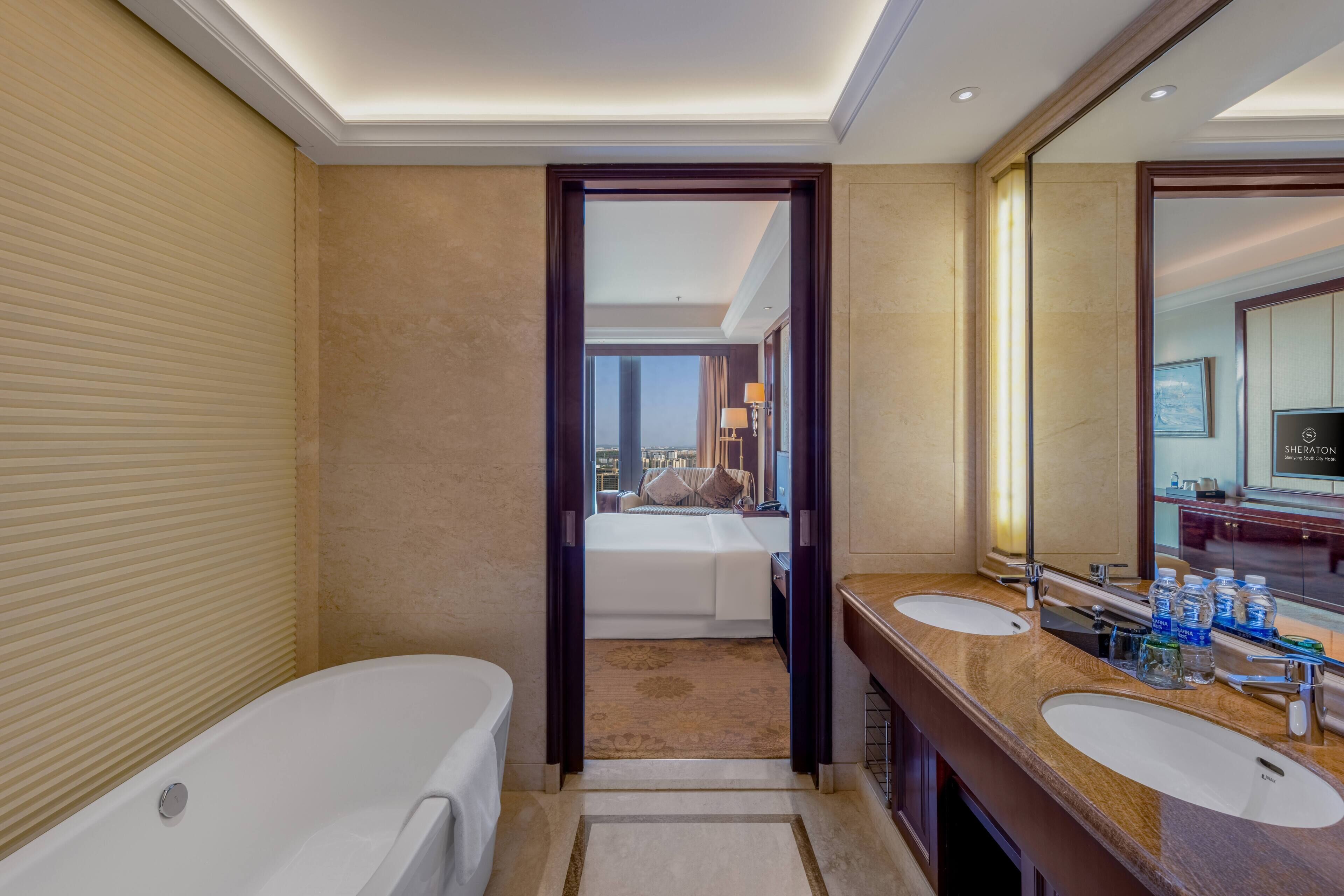 A hotel bathroom with a bathtub, two sinks, a large mirror, and a view of a bedroom with a bed and window.