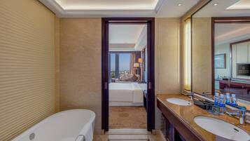 A hotel bathroom with a bathtub, two sinks, a large mirror, and a view of a bedroom with a bed and window.
