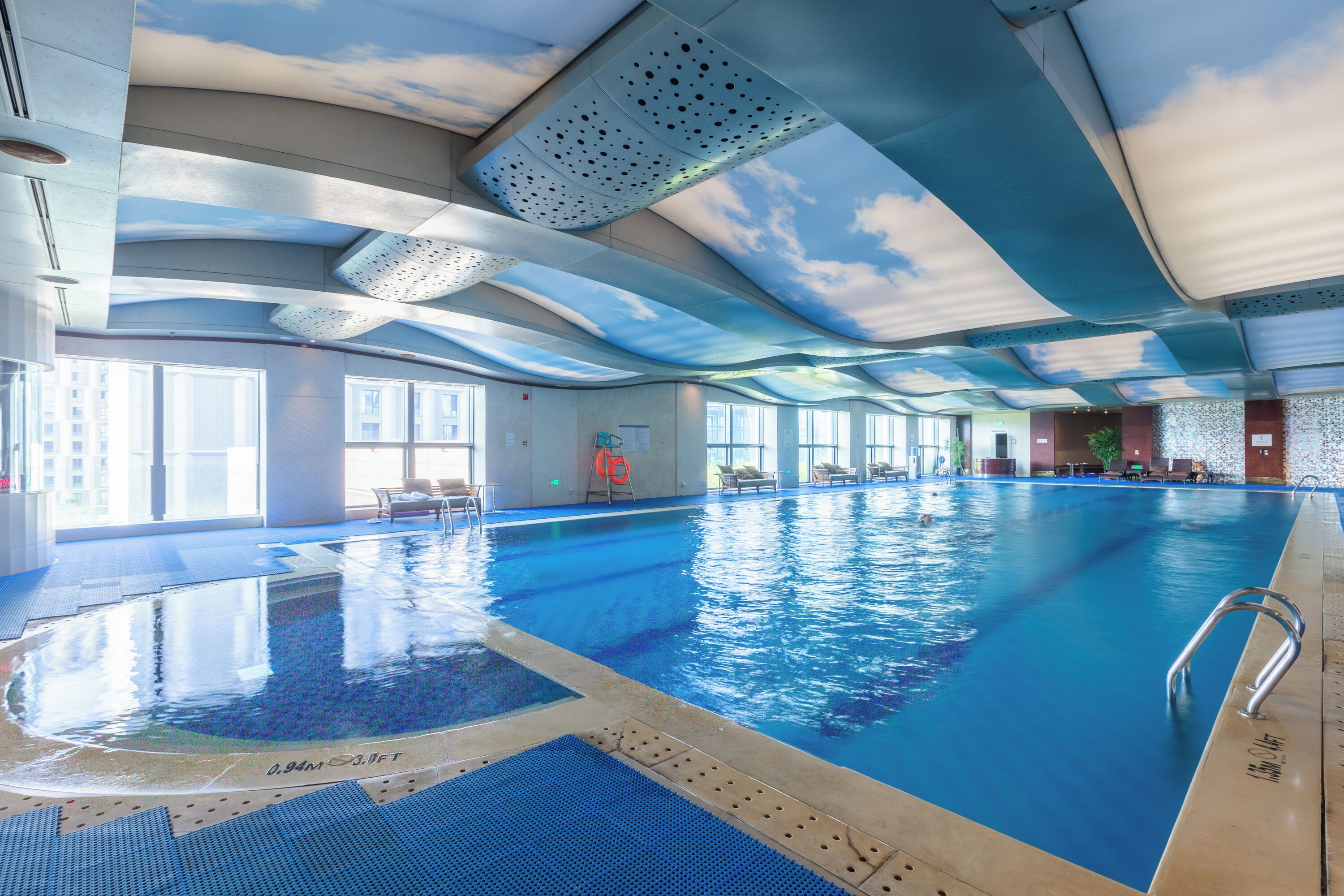 Una piscina interior con techo que simula un cielo nublado, ventanas amplias y una silla de salvavidas.
