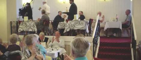 Un espace repas avec des tables et des chaises, une scène où se trouve un pianiste, et un escalier recouvert d’une moquette rouge.