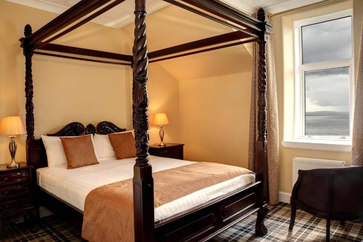 A four-poster bed with a wooden frame, a bedside table with a lamp, and a view of the sea through a window.