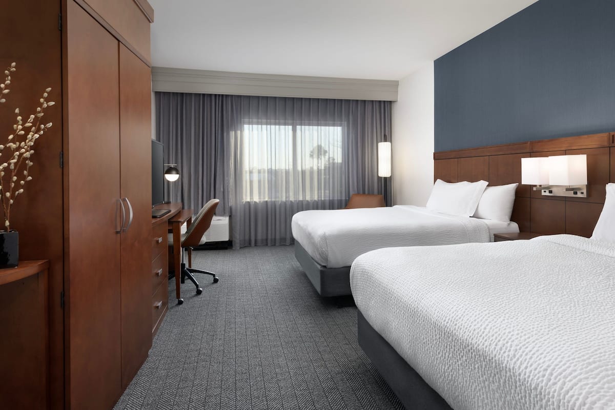 Ein Hotelzimmer mit zwei Betten, einem Schreibtisch, einem Stuhl, einem Kleiderschrank und einem großen Fenster mit Vorhängen.