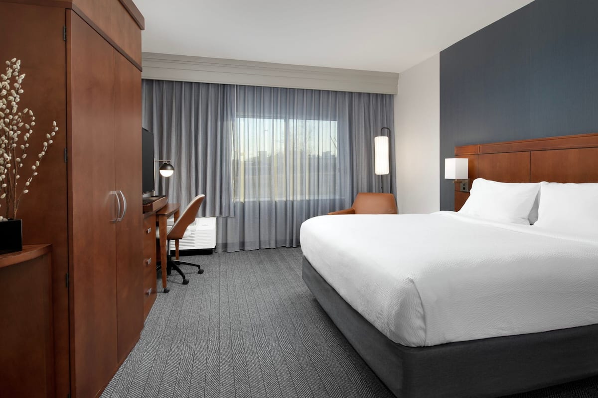 Ein Hotelzimmer mit einem großen Bett, einem Schreibtisch, einem Sessel und einem Kleiderschrank, mit Blick ins Freie.